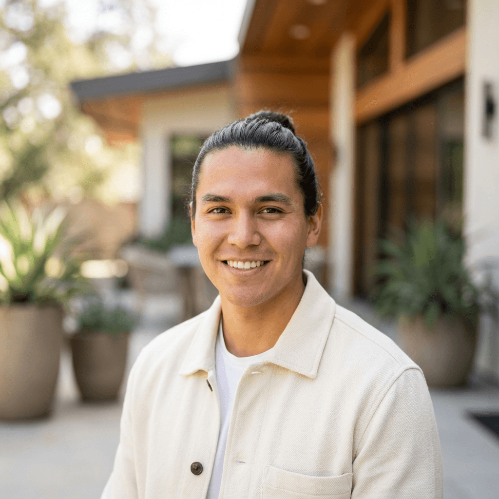 Smiling man with dark hair in a bun wearing a cream jacket on a patio.