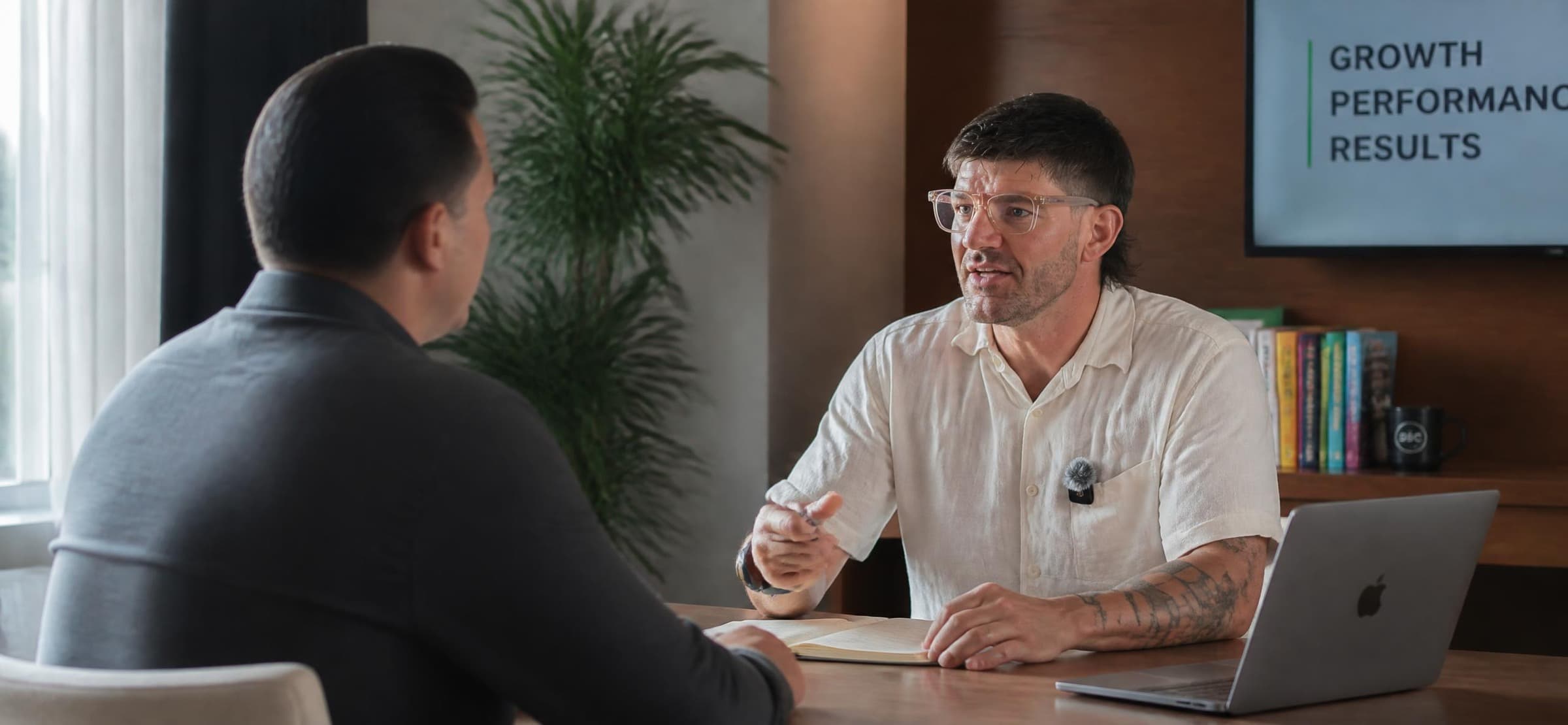 Man with glasses and tattoos gestures while speaking to a colleague in a professional office.