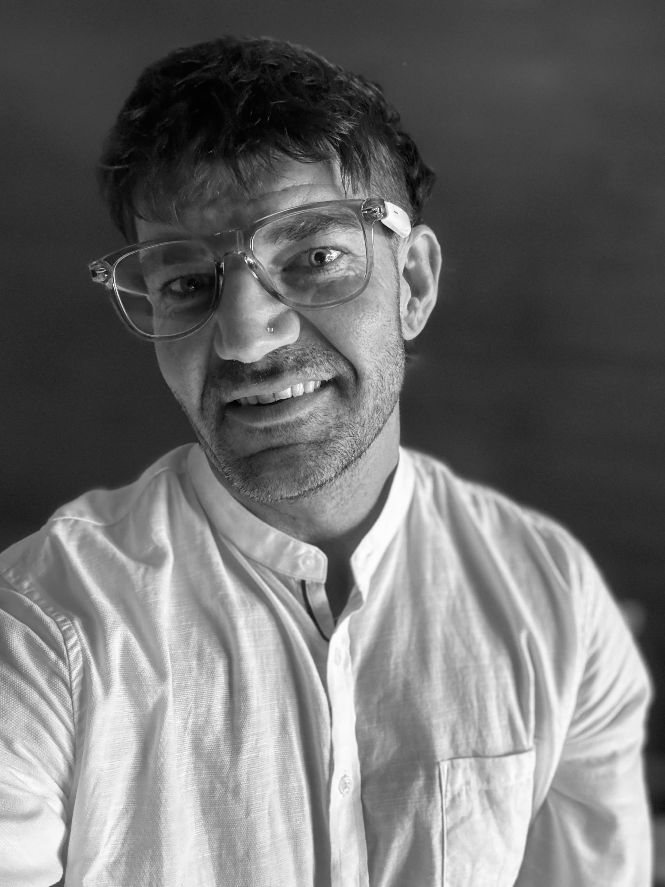 Monochrome portrait of a man with clear glasses and stubble wearing a white shirt.
